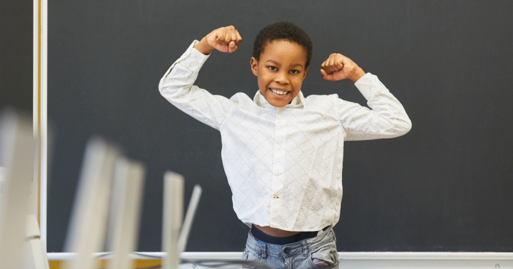 Young black student holding up his arms in a muscle formation with a smile. Happy and confident with a blackboard behind him in a classroom.