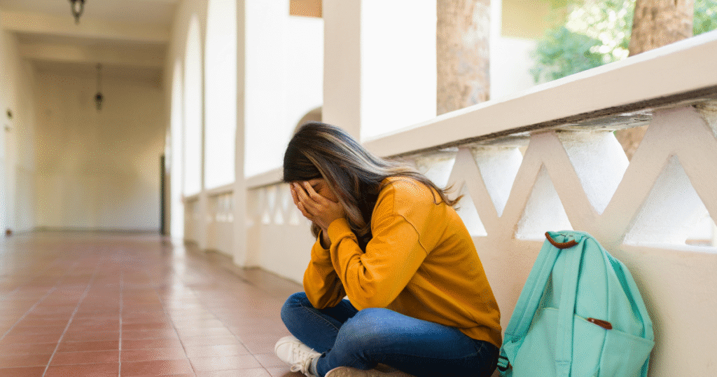 Young student in yellow shirt with teal backpack. Face in hands struggling in hallway.