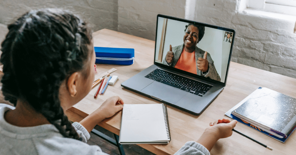 Black tutor on the laptop screen with thumbs up on a video call with student who has notebook and pen in front of her with the laptop.